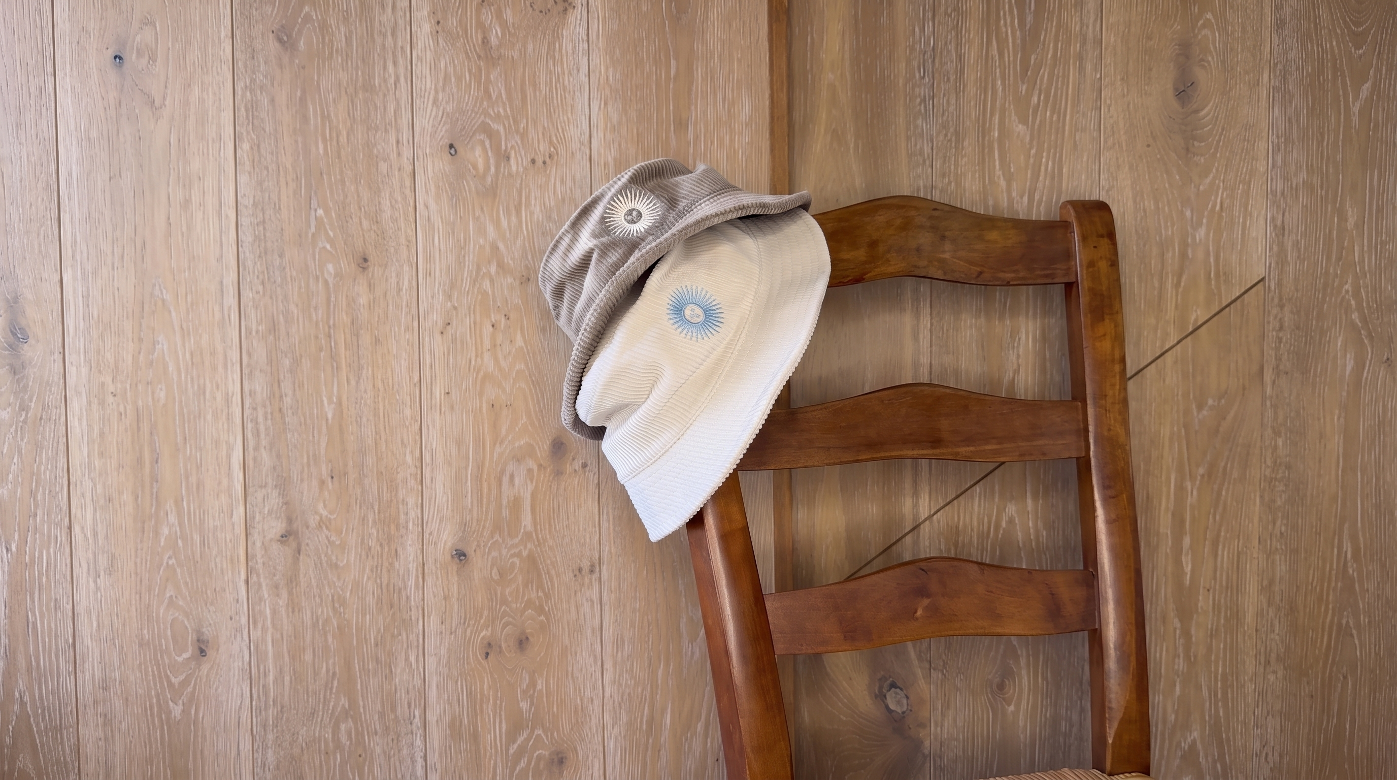 Two bucket hats, one beige and one off-white with a blue sunburst logo, draped over the back of a wooden chair against a wood-paneled wall.
