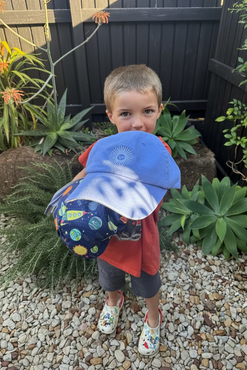 A young boy standing in a backyard garden setting, holding up a blue sun hat and a matching blue sun shield with a colorful space-themed pattern.