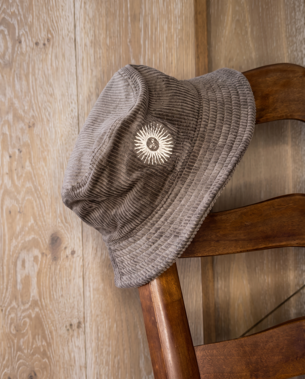 A taupe corduroy bucket hat with a white sunburst logo patch, resting on a rustic wooden chair against a light wood plank wall.