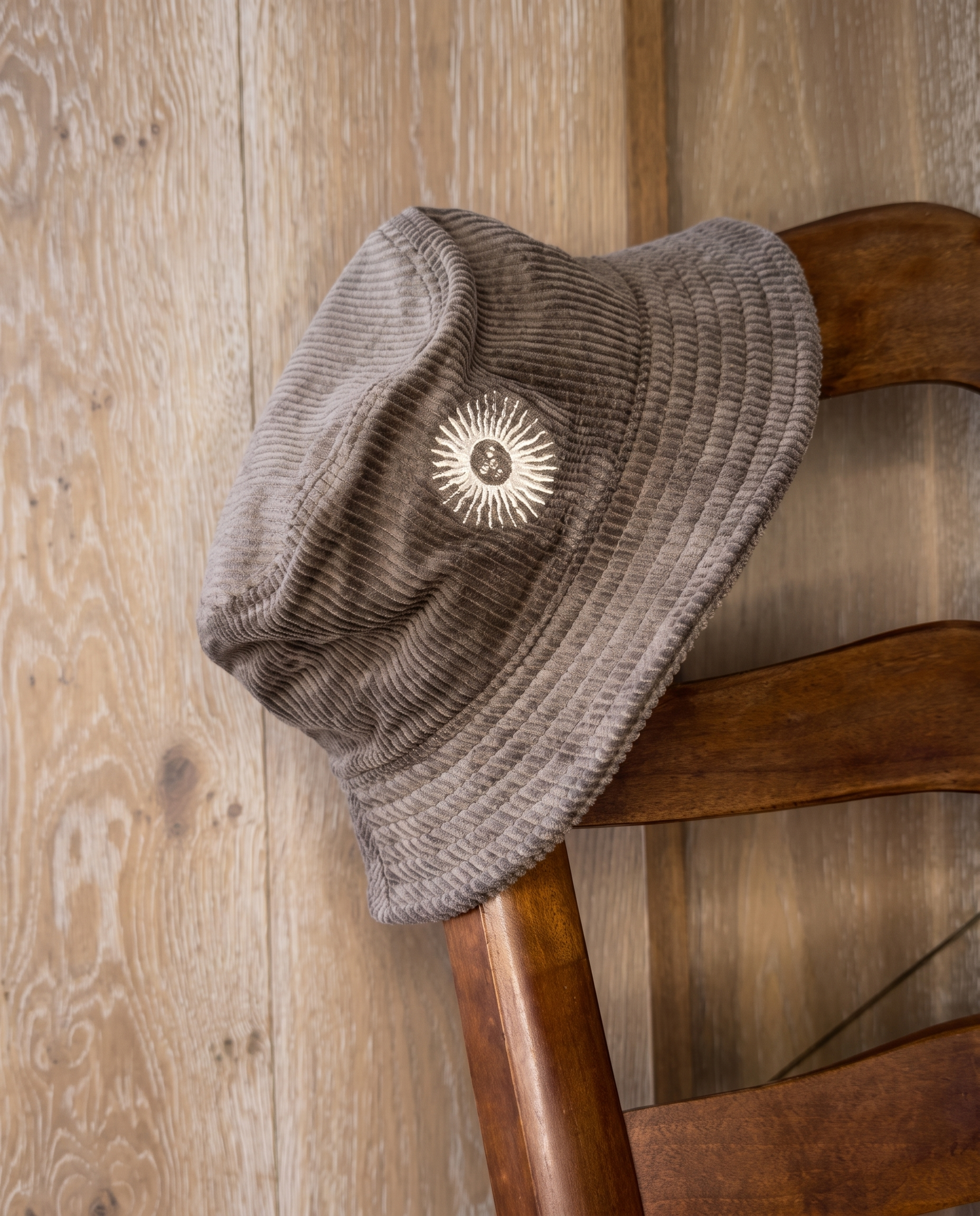 A taupe corduroy bucket hat with a white sunburst logo patch, resting on a rustic wooden chair against a light wood plank wall.