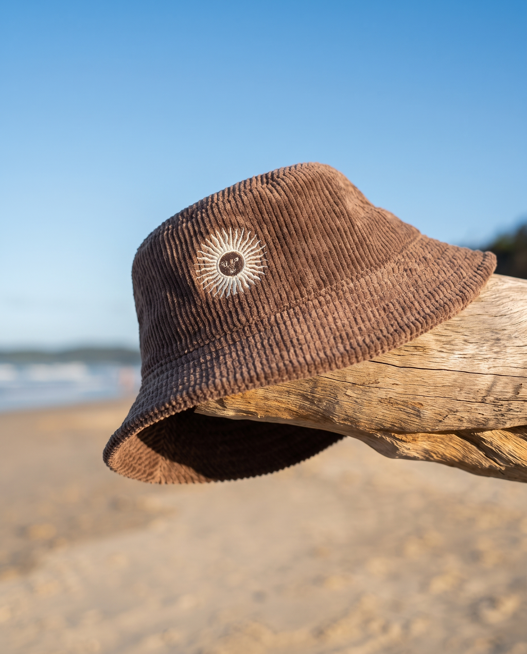 A brown corduroy bucket hat with a white sunburst logo on the front, resting on a piece of driftwood on a sandy beach with the ocean and blue sky in the background.