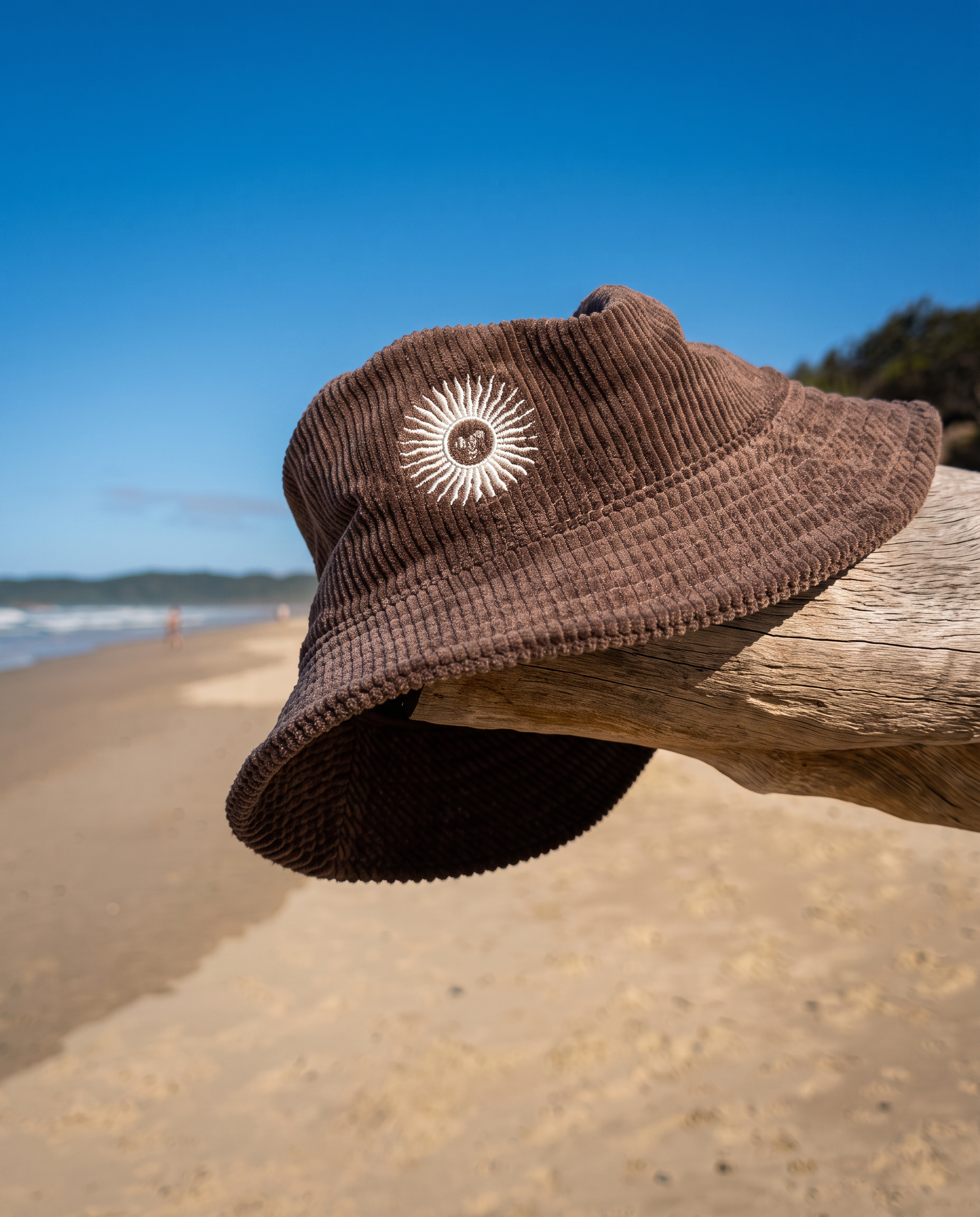 A brown corduroy bucket hat with a white sunburst logo sits on a piece of driftwood at a beach, with sand, ocean, and blue sky in the background.