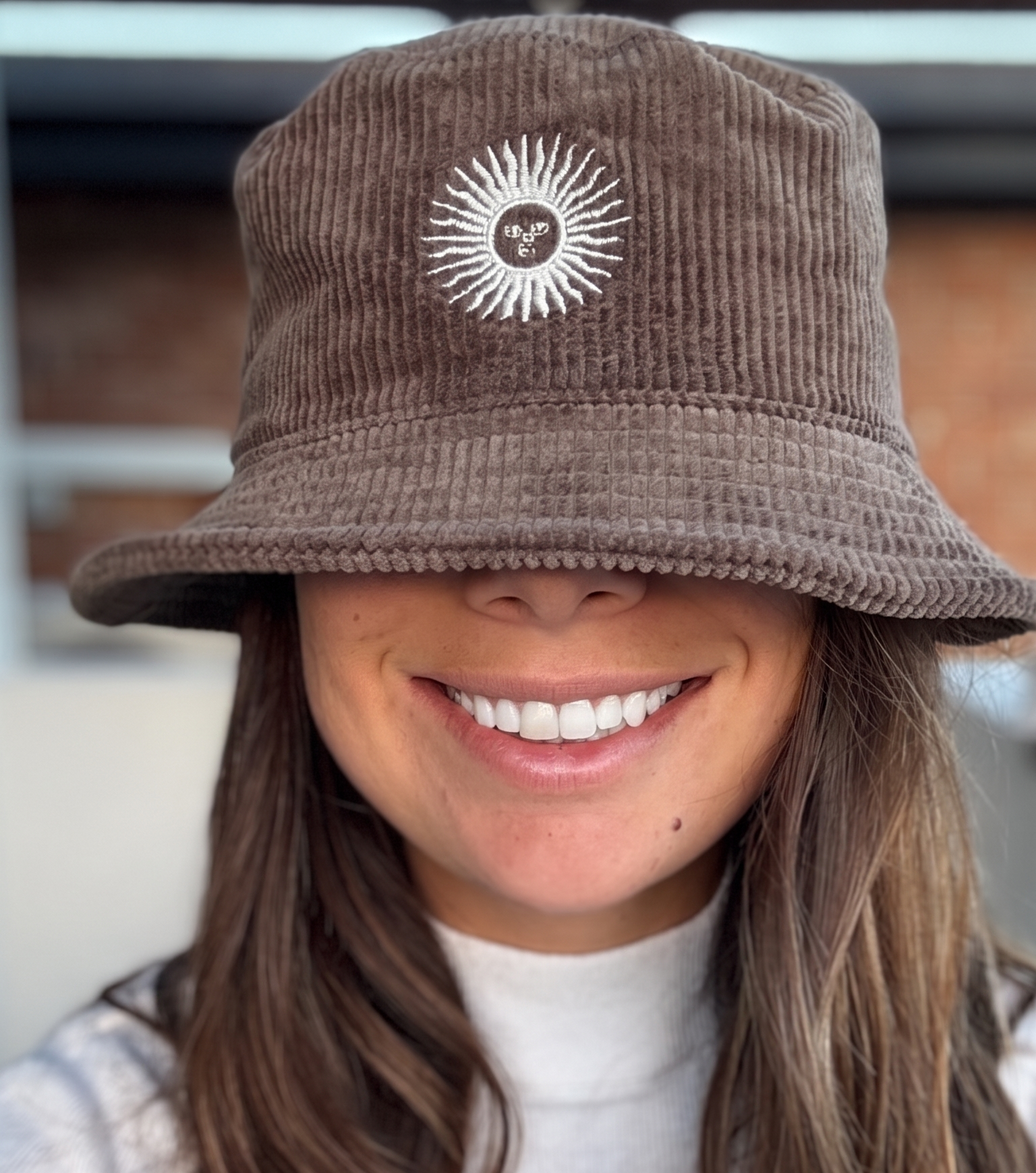 A woman wearing a brown corduroy bucket hat with a white embroidered sunburst logo on the front, photographed in a close-up selfie-style shot.